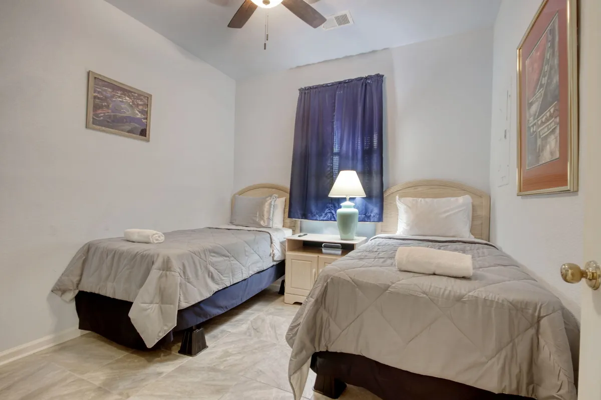 A peaceful twin bedroom featuring two neatly made beds, a decorative lamp, a window with dark blue curtains, and framed artwork. The setup suggests a quiet retreat perfect for unwinding after a long day.