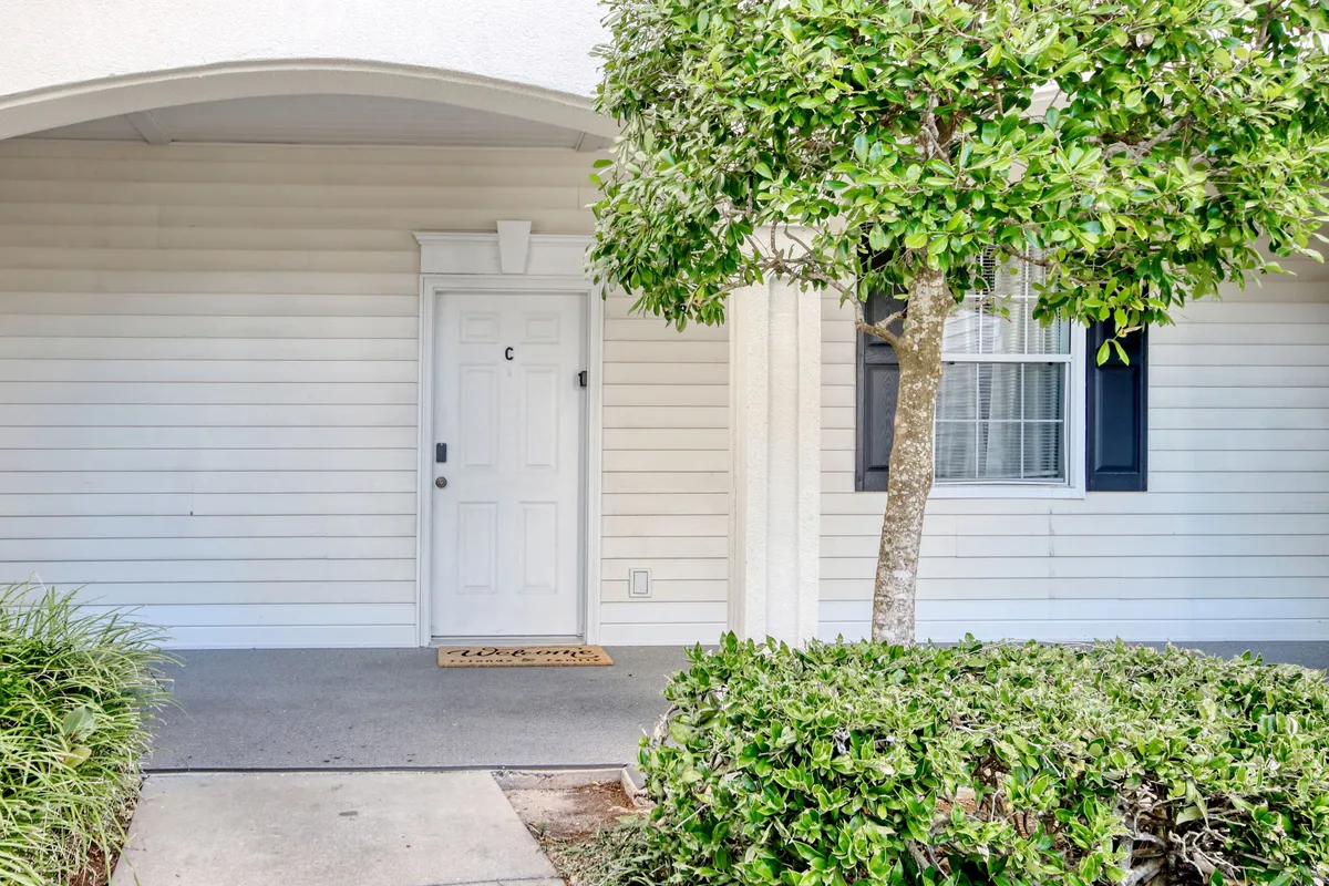 The front entrance to the condo, framed by a welcome mat, greenery, and a tree, suggesting a warm, inviting arrival point. The space conveys a sense of homecoming and a friendly neighborhood ambiance.