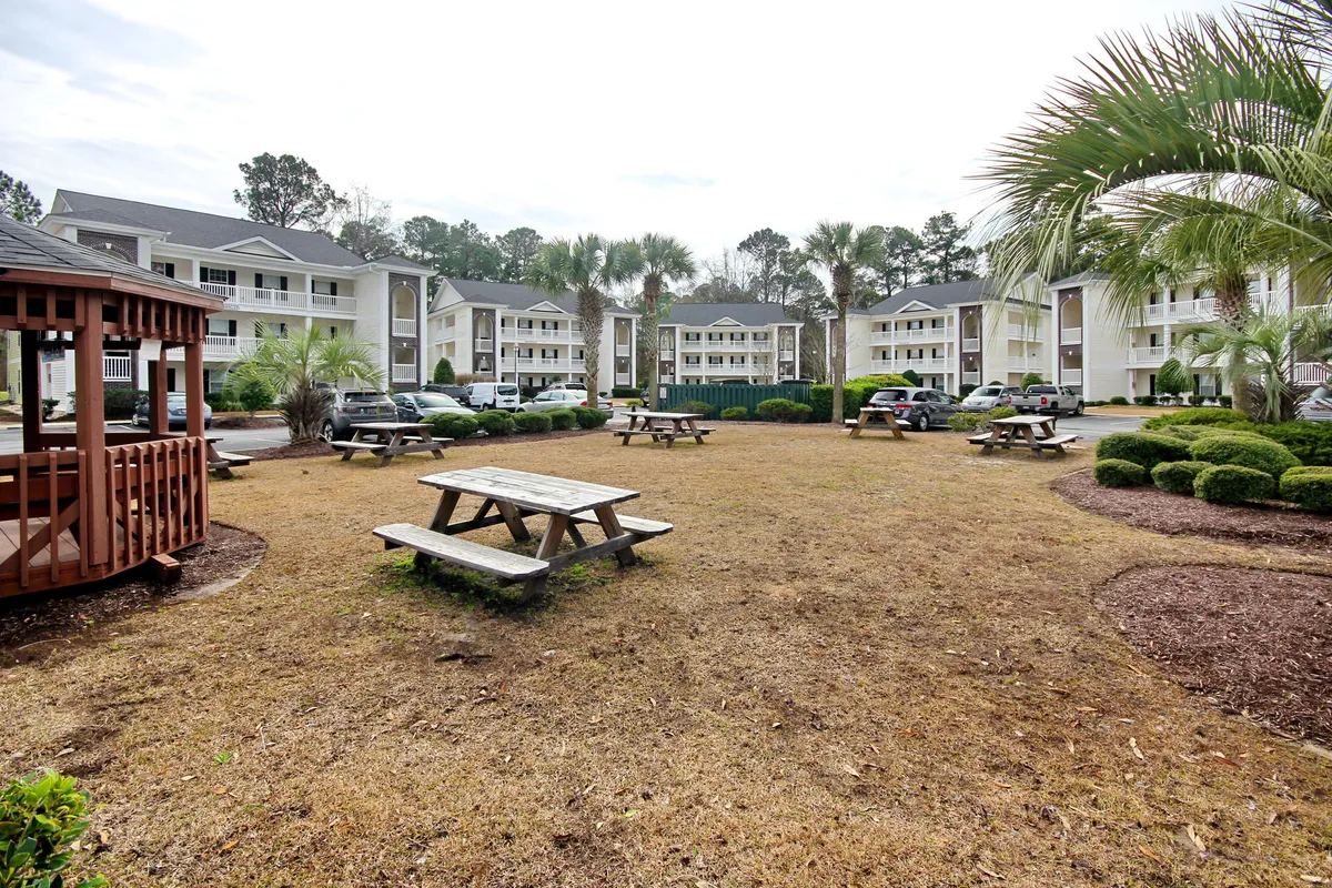 A cheerful outdoor space featuring picnic tables beneath a gazebo, with manicured bushes and nearby apartment buildings. The space is designed for gatherings, outdoor meals, and playful activities, inviting families to enjoy fresh air and togetherness.