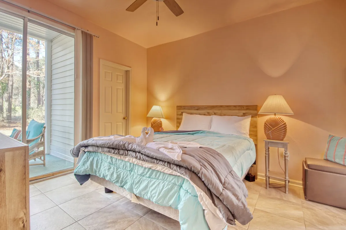 This image showcases a serene bedroom featuring a bed with a light blue comforter, a large window framing a view of the outdoors, and decorative lamps that cast a warm, tranquil glow. The soft, calming décor and plush bedding immediately evoke a sense of relaxation and comfort.