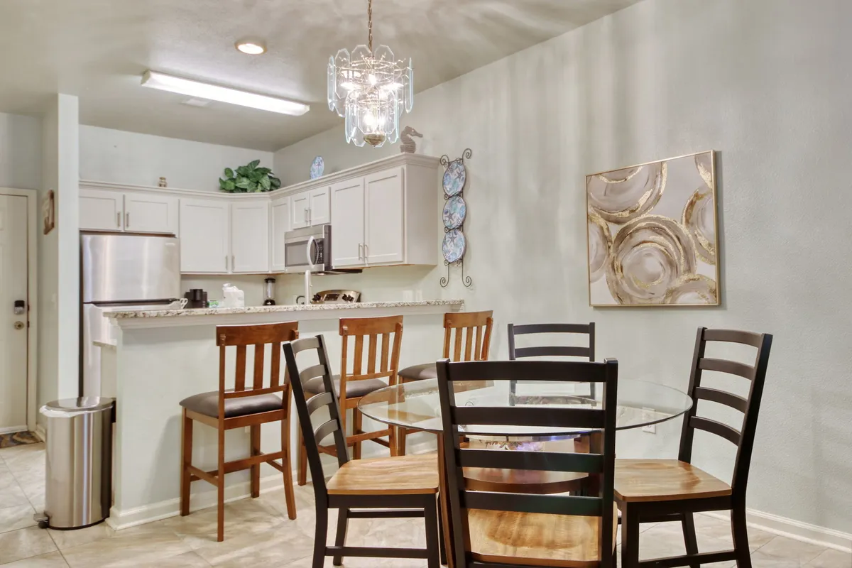 An inviting dining area with a glass dining table, wooden chairs, a stylish chandelier overhead, and a glimpse of the kitchen with an island and barstools. Decorative plates adorn the wall, contributing to a homey and welcoming atmosphere ideal for family meals and celebrations.