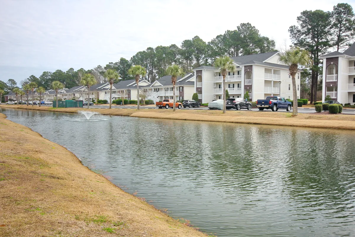 A serene pond with a fountain surrounded by palm trees and residential buildings. The calm water reflects the blue sky, and the landscaping suggests a peaceful, community-oriented environment ideal for leisurely walks after a day of golf.